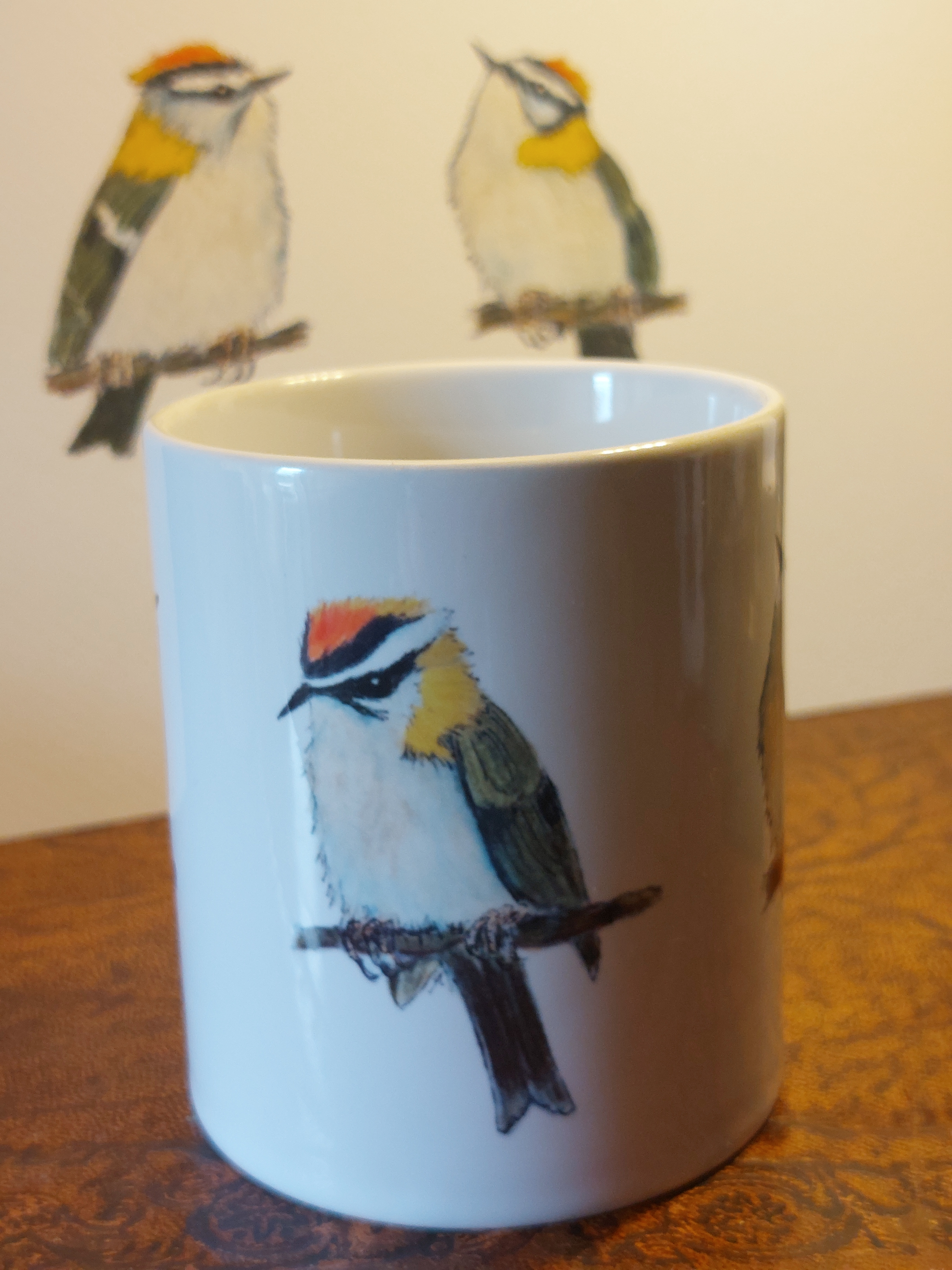 Ceramic mug with Firecrest painting by fairy flint printed with eco friendly inks, mug on amtique wooden shelf in front of print of Firecrests