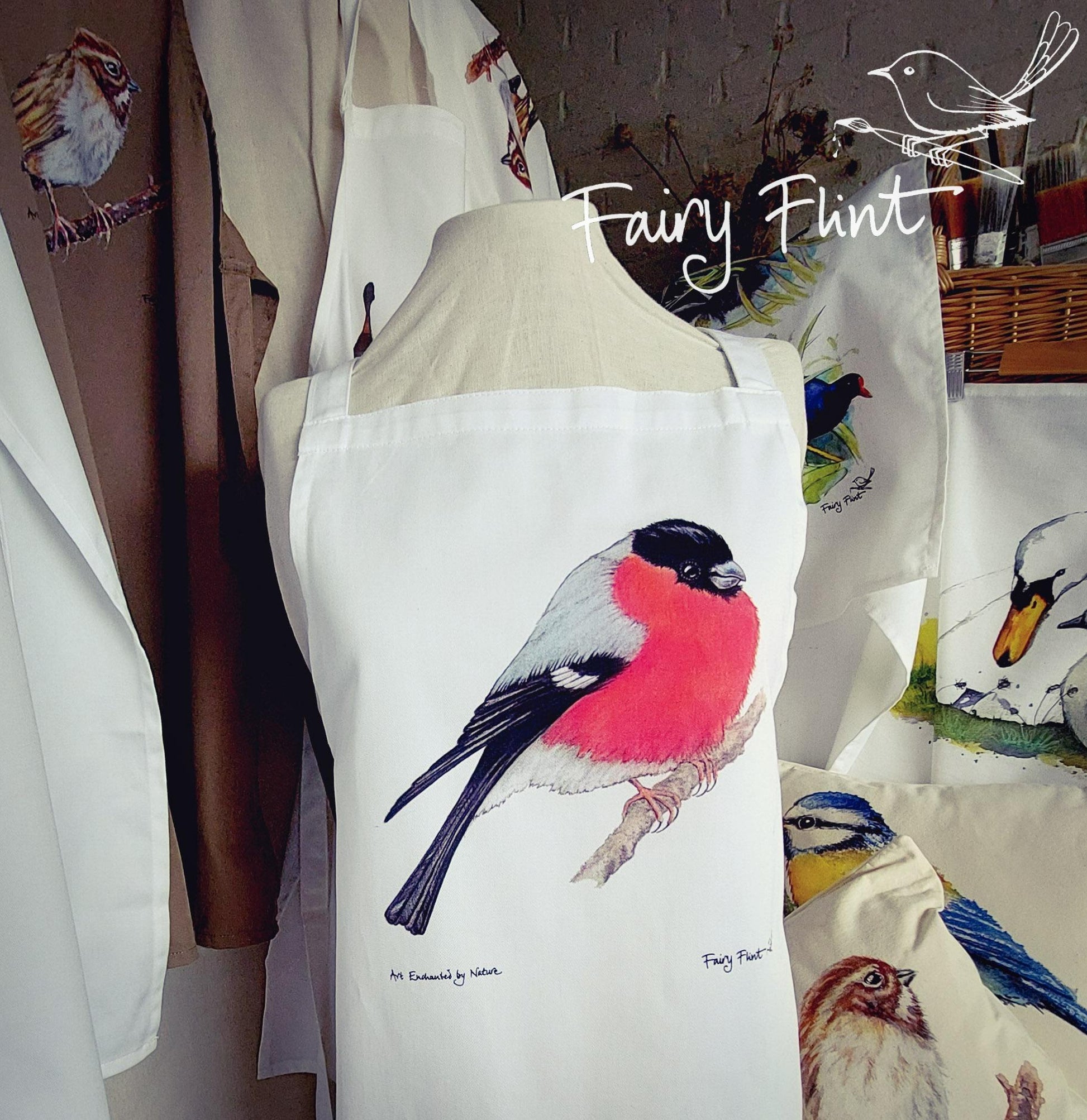 Organic Apron with Bullfinch painting by Fairy Flint printed with eco friendly inks, apron hung on mannequin in front of other aprons with birds and ducks and swans on