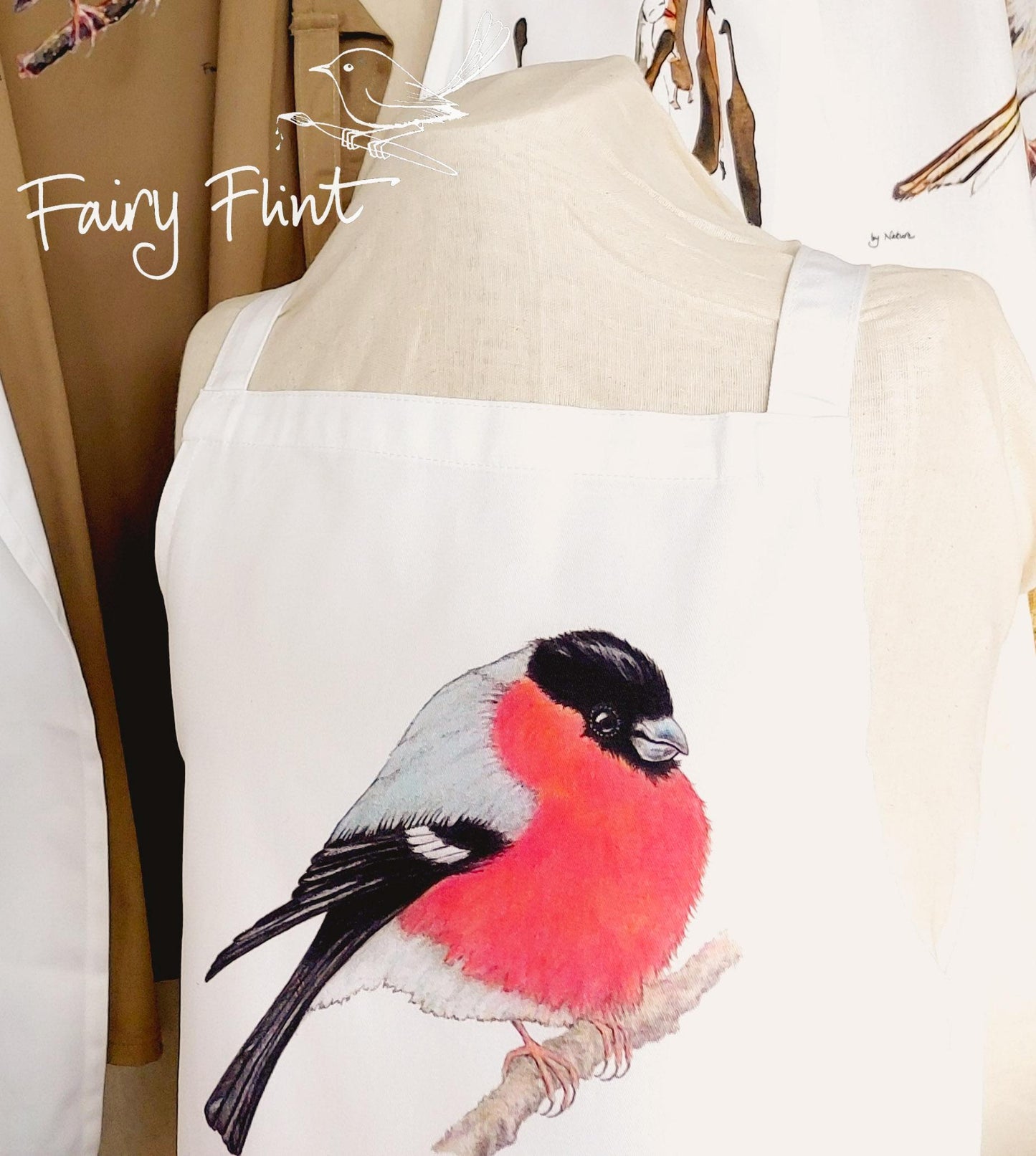 Organic Apron with Bullfinch painting by Fairy Flint printed with eco friendly inks, apron hung on mannequin in front of other aprons with birds and ducks on