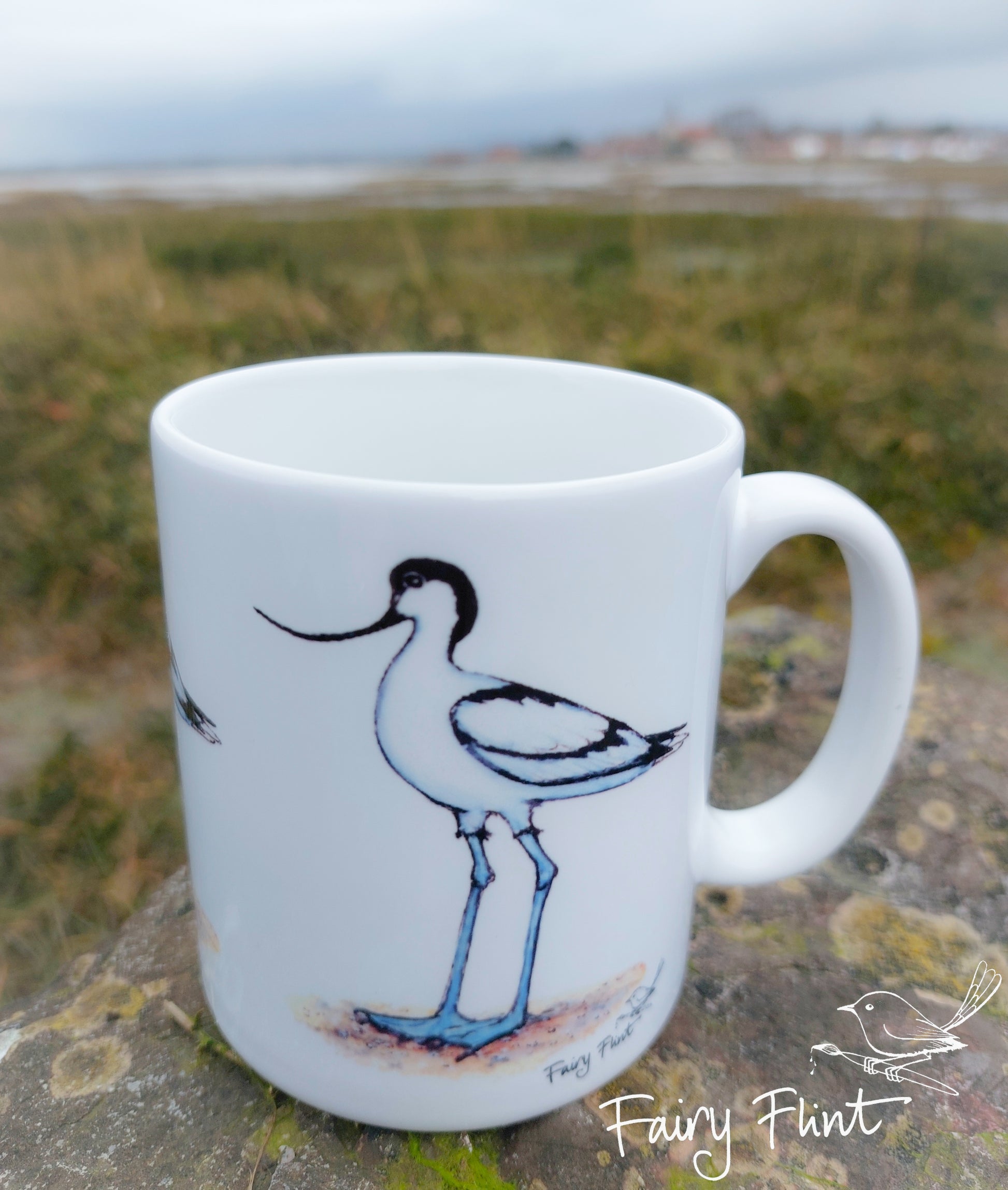 Ceramic mug with Avocet painting eco friendly print by Fairy Flint, photoshoot at Bosham harbour, mug standing on lichen covered rock in front of harbour and sea in background