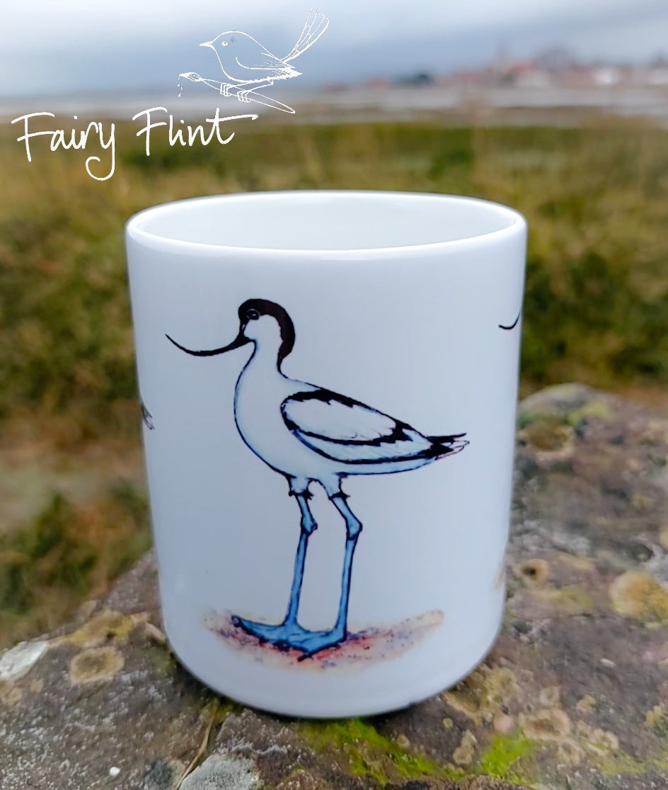 Ceramic mug with Avocet painting eco friendly print by Fairy Flint, photoshoot at Bosham harbour, mug standing on lichen covered rock in front of harbour and sea in background