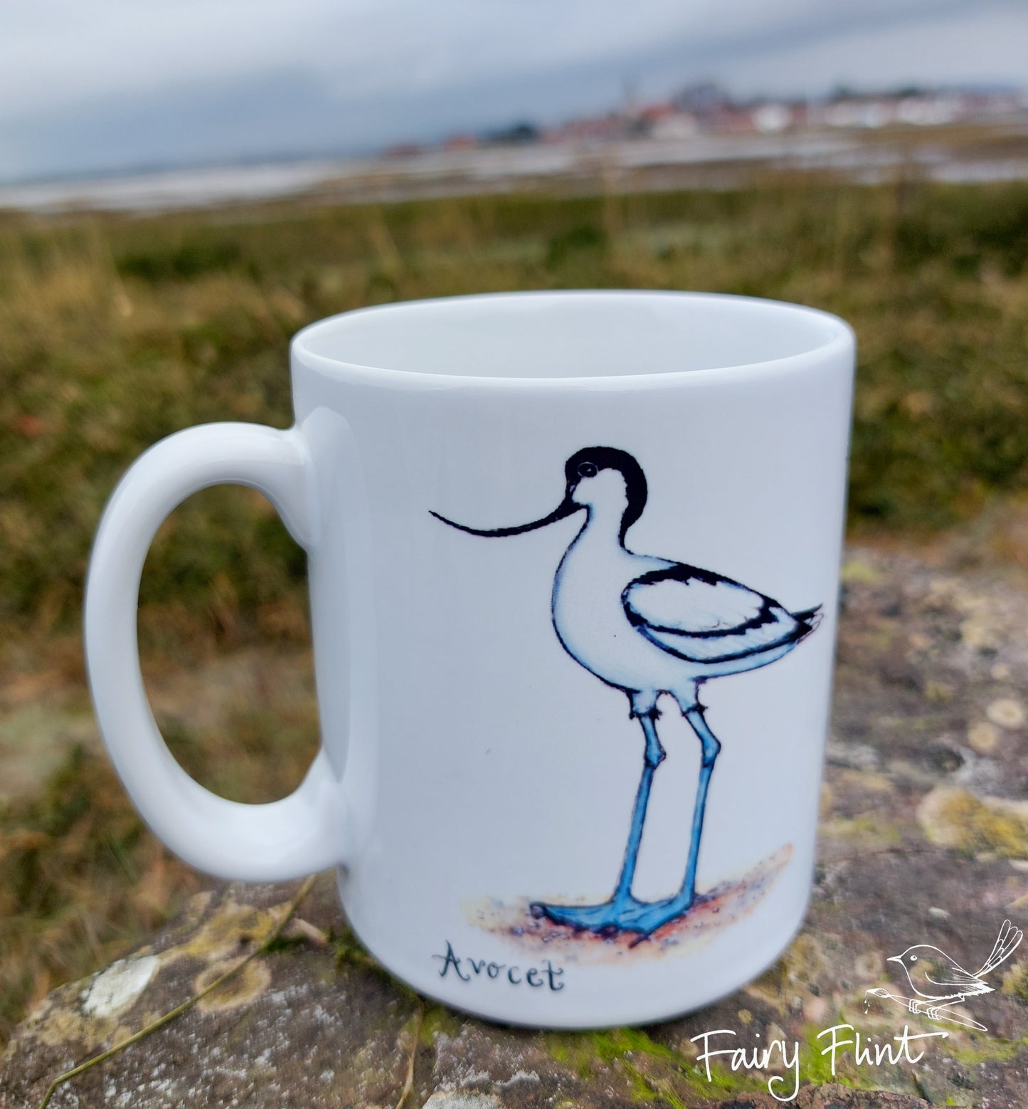 Ceramic mug with Avocet painting eco friendly print by Fairy Flint, photoshoot at Bosham harbour, mug standing on lichen covered rock in front of harbour and sea in background