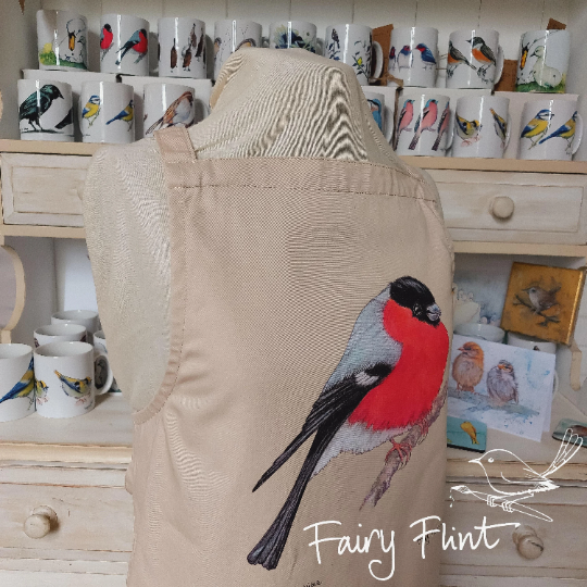 Organic Apron with Bullfinch painting by Fairy Flint printed with eco friendly inks, apron hung on mannequin in front of other aprons with birds, moorhens, ducks and swans on