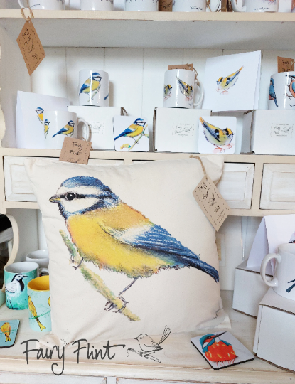 Canvas natural cushion with eco friendly ink print of Blue tit by Fairy Flint, cushion on dresser with other blue tit mugs coaster and goldcrest coaster behind behind, wagtail mug on shelf and kingfisher coaster 